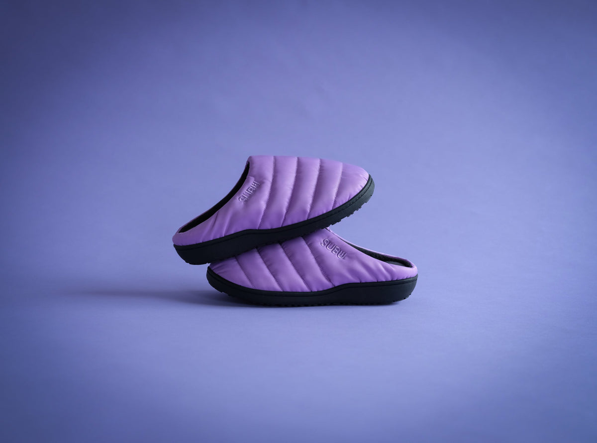 A pair of lavender purple quilted slip-on slippers with black soles, stacked on a matching purple studio background.
